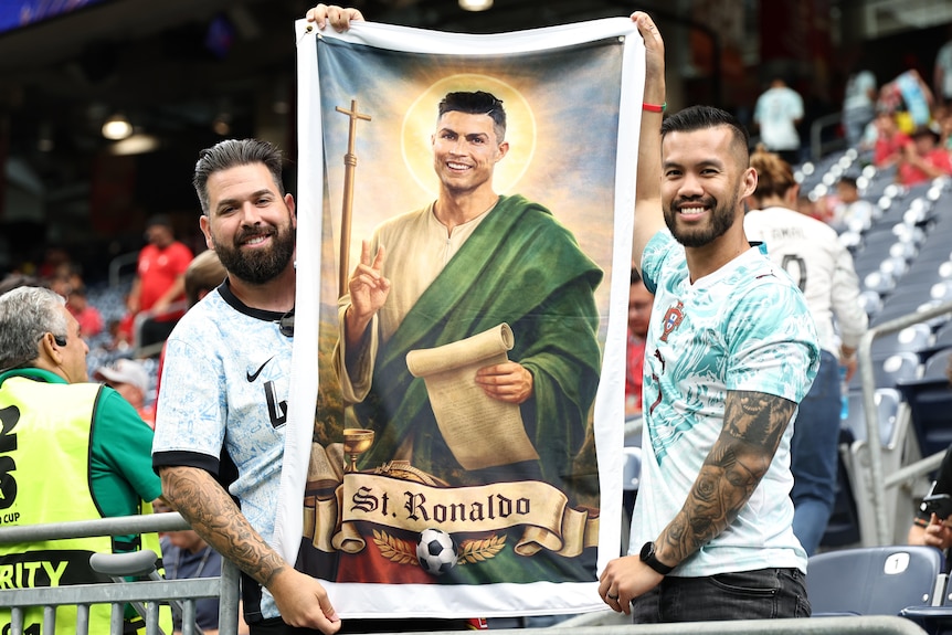 Two Portugal fans inside a stadium hold a banner which depicts Cristiano Ronaldo as a saint.